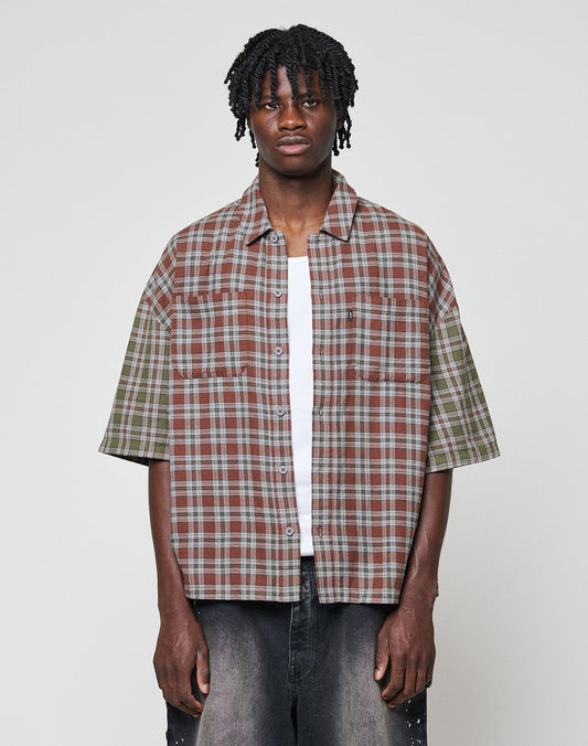 A young man with short dreadlocks wears the LIVE FAST DIE YOUNG LFDY Light Flannel—an oversize fit, short-sleeve, plaid button-up in brown, green, and grey—over a white tee and dark jeans against a plain light background. | model