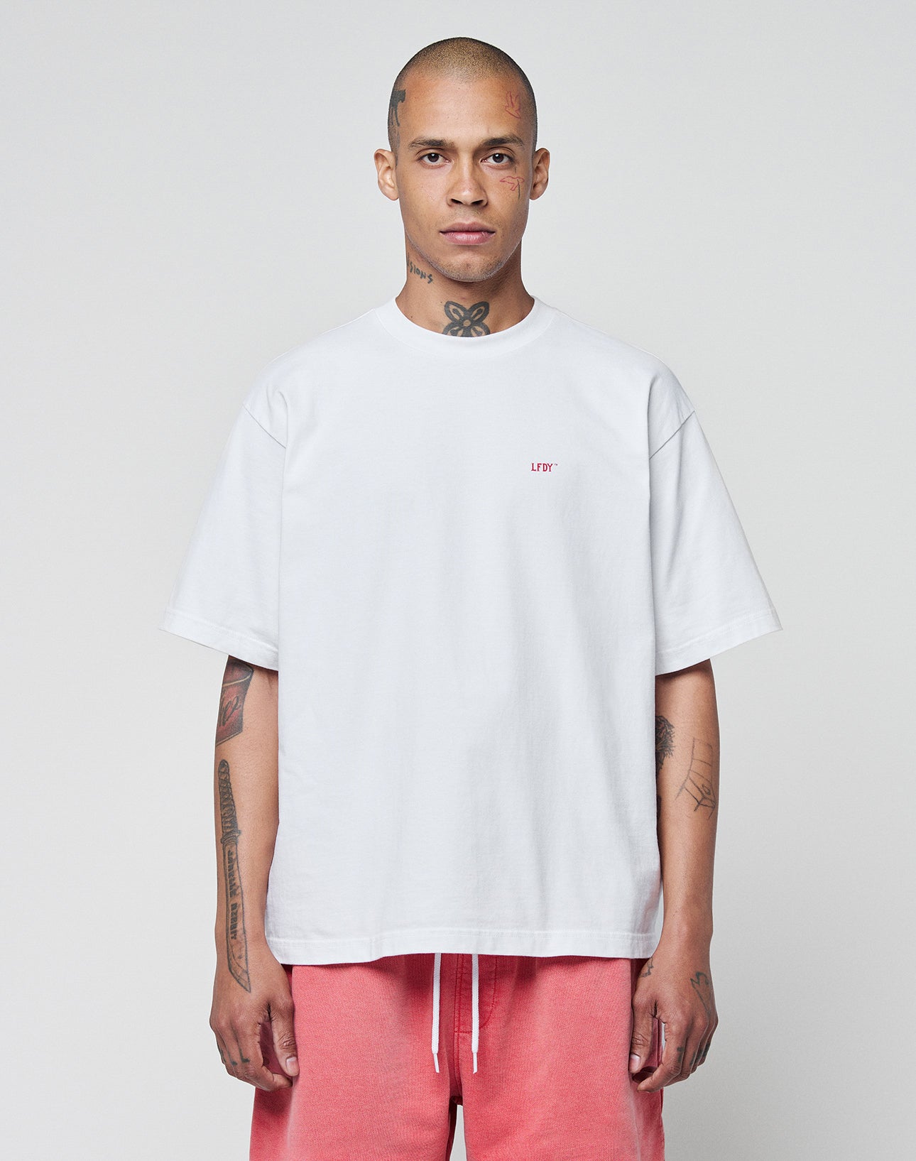 A person with a shaved head and tattoos faces forward, wearing the LIVE FAST DIE YOUNG Basic Evidence Tee in white with small red “LBY” lettering, paired with pink drawstring shorts, set against a plain light background.