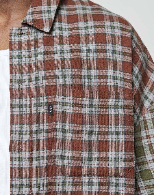Close-up of a person wearing the LFDY Light Flannel by LIVE FAST DIE YOUNG in an oversize, brown, white, and green plaid design with vintage wash and a black LFDY tag on the chest pocket. Only part of the chest and shoulder is visible.