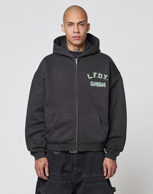 A man with a shaved head wears the Live Fast District Hooded Zip by LIVE FAST DIE YOUNG, an oversized black zip-up jacket with light blue LFDY branding on the chest, standing against a plain white background.