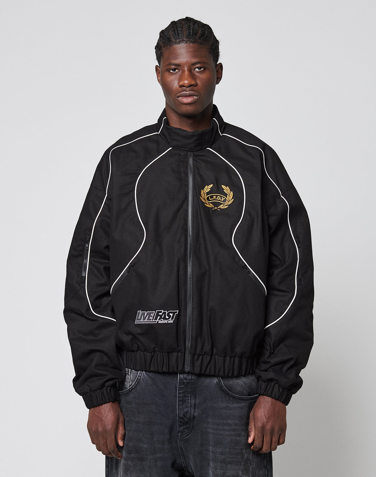 A man with braided hair wears the LFDY Canvas Jacket by LIVE FAST DIE YOUNG, featuring white piping, a gold chest crest, multi-logo patches, MEH.T text near the hem, and elastic cuffs against a plain light gray background.