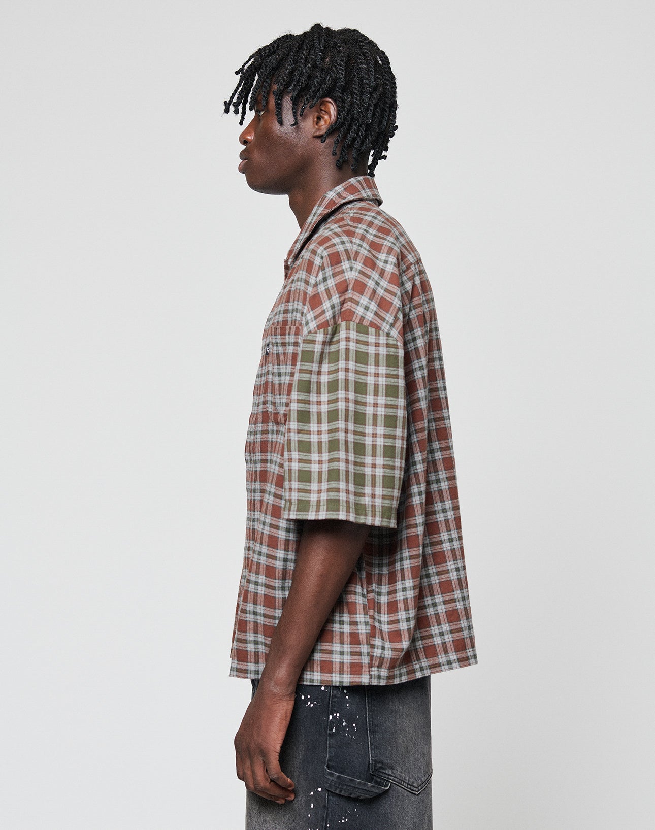 A person with short, twisted hair models the LIVE FAST DIE YOUNG LFDY Light Flannel—an oversize fit, short-sleeve plaid shirt with mismatched brown and green sleeves—paired with dark jeans against a plain light background.