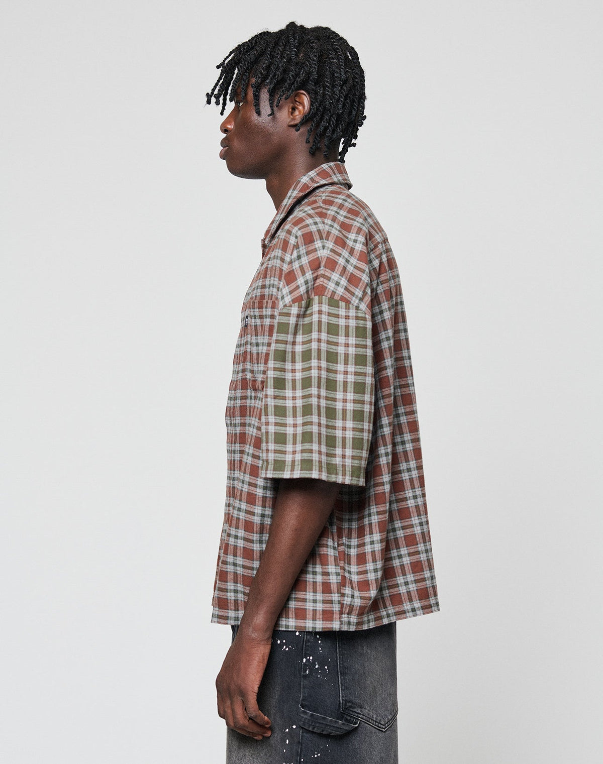 A person with short, twisted hair models the LIVE FAST DIE YOUNG LFDY Light Flannel—an oversize fit, short-sleeve plaid shirt with mismatched brown and green sleeves—paired with dark jeans against a plain light background.