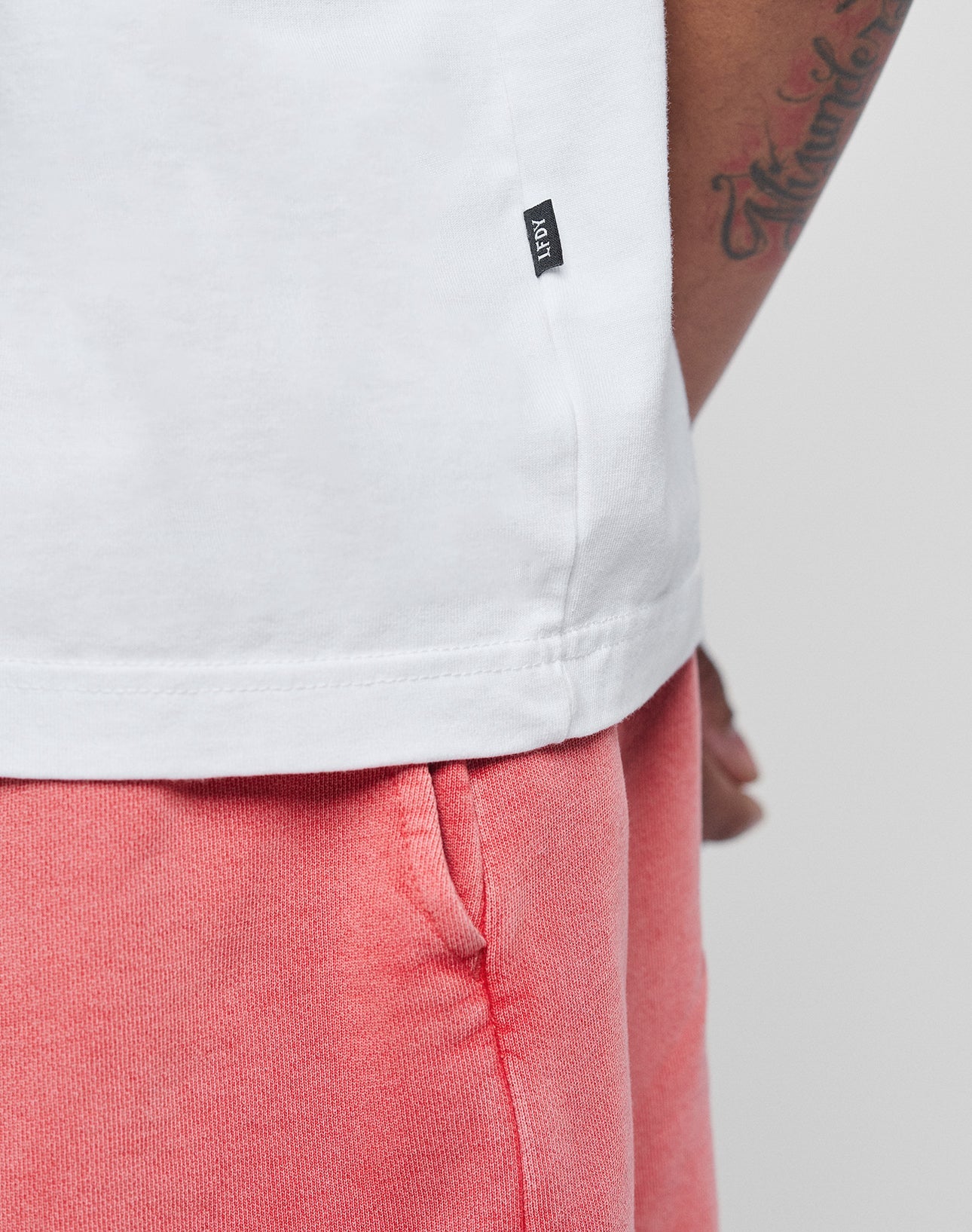 Close-up of a person wearing the LIVE FAST DIE YOUNG Basic Evidence Tee in white, featuring a small black tag, paired with pink shorts. The soft tee is crafted from 100% Baumwolle for comfort and style; tattooed arm and pocket detail visible.