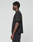 A person with short curly hair stands in profile against a plain background, wearing the LIVE FAST DIE YOUNG Basic 210 Tee in black. The oversize fit t-shirt features front and back graphics and text on the sleeve, paired with black pants.