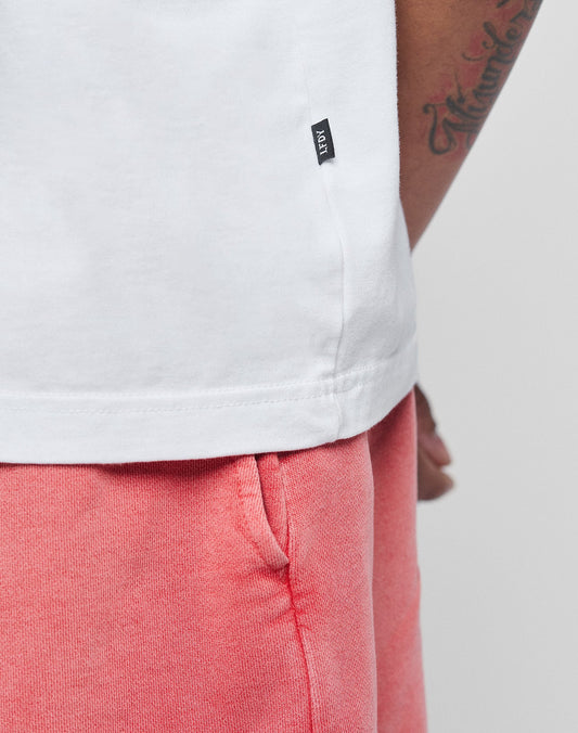 Close-up of a person wearing the LIVE FAST DIE YOUNG Basic Evidence Tee in white, featuring a small black tag, paired with pink shorts. The soft tee is crafted from 100% Baumwolle for comfort and style; tattooed arm and pocket detail visible.