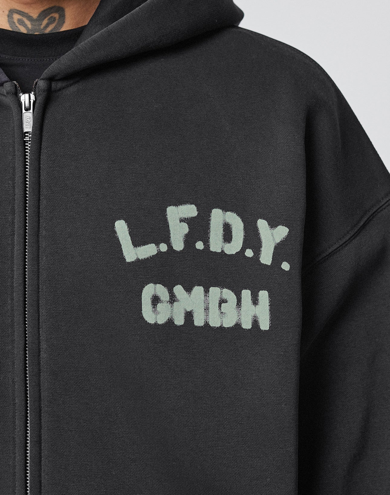 Someone wears the Live Fast District Hooded Zip by LIVE FAST DIE YOUNG, a black oversized zip-up hoodie with “L.F.D.Y. GMBH” in pale, blocky letters on the left chest; a neck tattoo peeks above the collar for a bold finish.