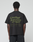 A person stands with their back to the camera, wearing the LIVE FAST DIE YOUNG Basic 210 Tee—an oversize black T-shirt made from 100% cotton, featuring olive green “LiveFast ArmyGrn 17-0517” text on the back, against a plain white background.