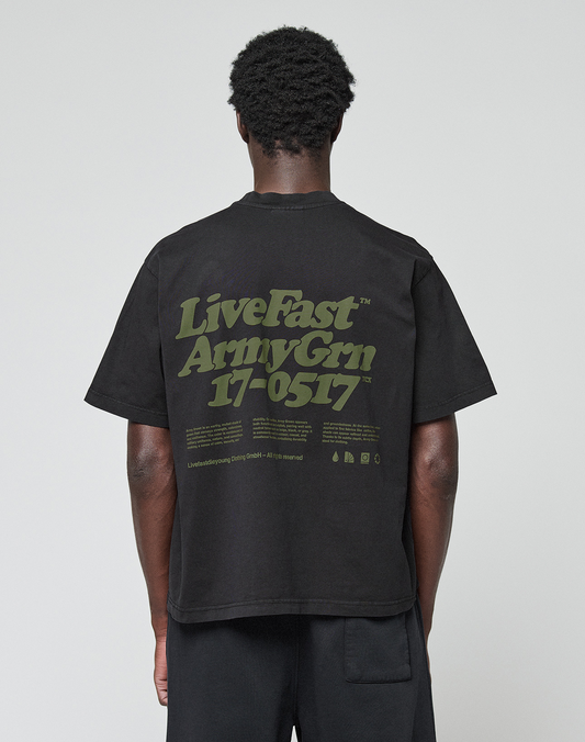 A person stands with their back to the camera, wearing the LIVE FAST DIE YOUNG Basic 210 Tee—an oversize black T-shirt made from 100% cotton, featuring olive green “LiveFast ArmyGrn 17-0517” text on the back, against a plain white background.