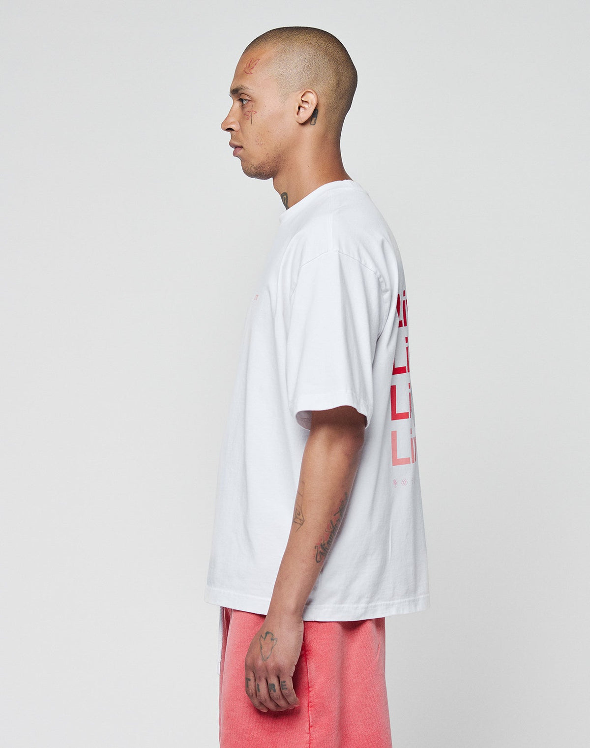 A tattooed person with a shaved head stands in profile against a plain background, wearing the LIVE FAST DIE YOUNG Basic Evidence Tee—an oversized white T-shirt aus 100% Baumwolle with red front and back print—and salmon pink shorts.