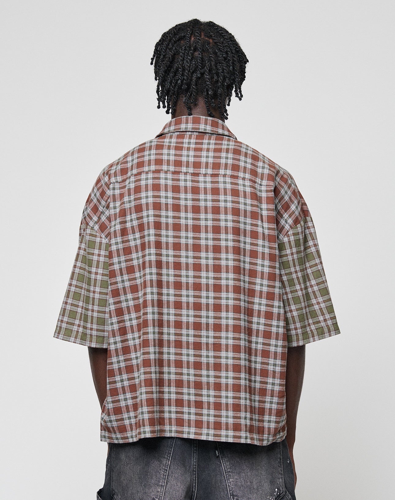 A person with short, twisted hair is seen from behind wearing the LIVE FAST DIE YOUNG LFDY Light Flannel—an oversized, short-sleeved plaid shirt in brown and green—paired with dark denim shorts against a plain background.