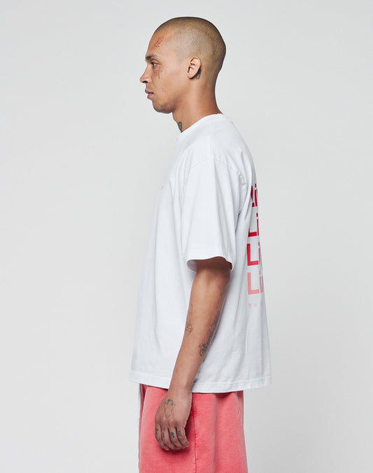 A tattooed person with a shaved head stands in profile against a plain background, wearing the LIVE FAST DIE YOUNG Basic Evidence Tee—an oversized white T-shirt aus 100% Baumwolle with red front and back print—and salmon pink shorts.