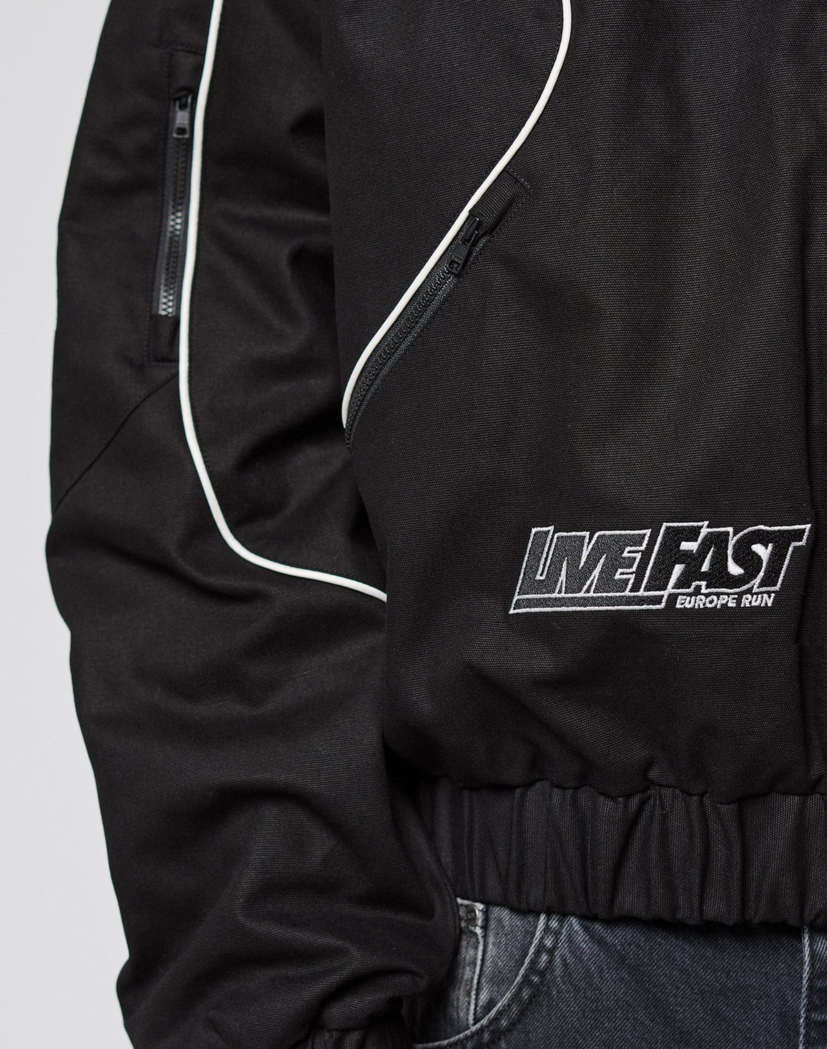 Close-up of a black LFDY Canvas Jacket by LIVE FAST DIE YOUNG with white piping, elastic cuffs, zippered pocket, and LIVE FAST EUROPE RUN embroidery near the waist. Shown paired with dark jeans.
