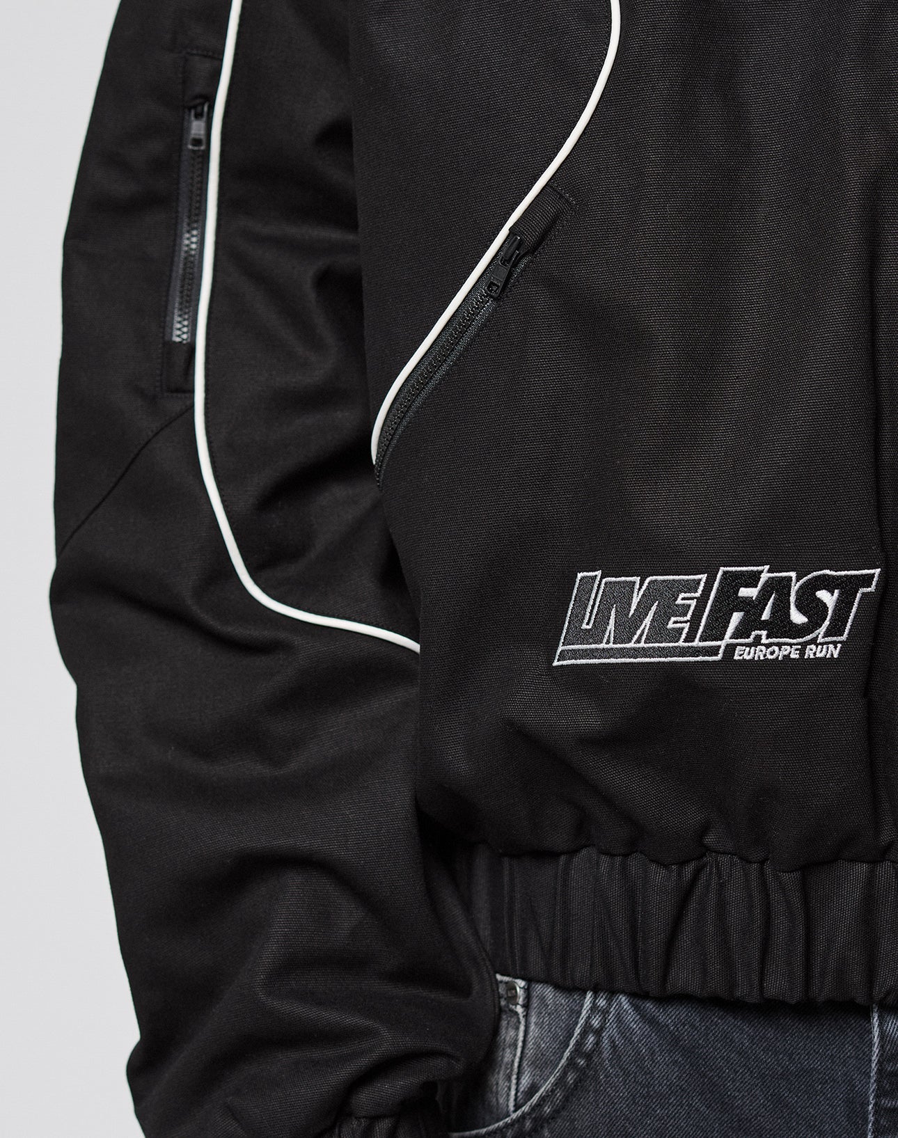 Close-up of a black LFDY Canvas Jacket by LIVE FAST DIE YOUNG with white piping, elastic cuffs, zippered pocket, and LIVE FAST EUROPE RUN embroidery near the waist. Shown paired with dark jeans.