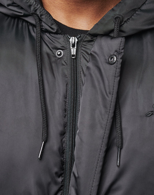 Close-up of a person in the LIVE FAST DIE YOUNG LFDY Winter Bomber Jacket, showing only the lower face, neck, and chest. The shiny black polyester features a hood with metal eyelets and toggles for an Oversize Fit look.