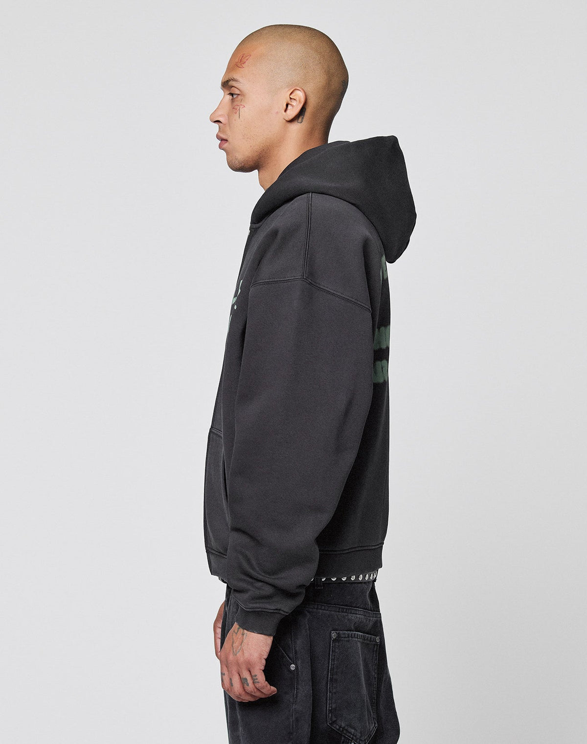 A person with a shaved head and visible tattoos stands in profile against a plain background, wearing the LIVE FAST DIE YOUNG Live Fast District Hooded Zip in black with Oversize Fit and black jeans, hands by their sides.