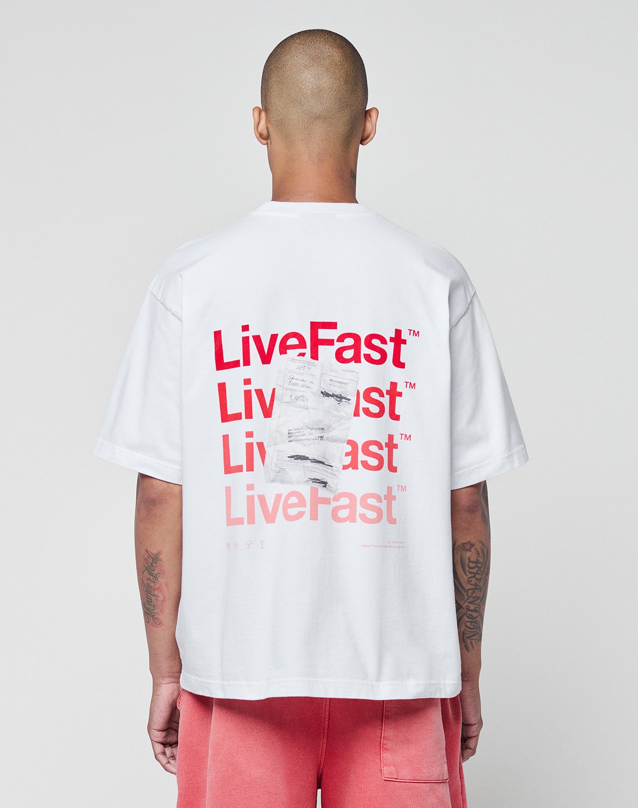 Seen from behind, a person with a shaved head wears the LIVE FAST DIE YOUNG Basic Evidence Tee—a white, 100% Baumwolle oversize T-shirt with red LiveFast prints on front and back—paired with pink shorts against a plain light background.