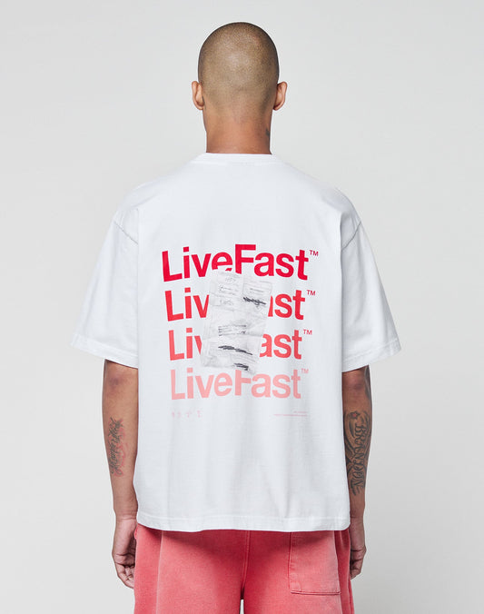 Seen from behind, a person with a shaved head wears the LIVE FAST DIE YOUNG Basic Evidence Tee—a white, 100% Baumwolle oversize T-shirt with red LiveFast prints on front and back—paired with pink shorts against a plain light background. | model