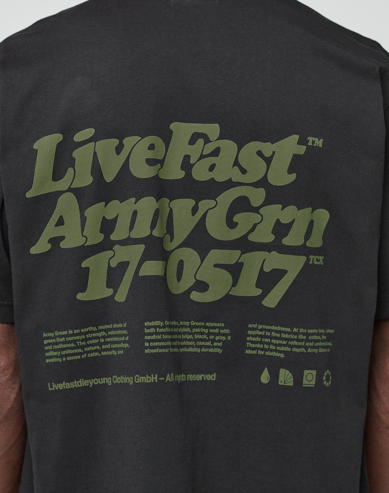 A close-up of the Basic 210 Tee by LIVE FAST DIE YOUNG shows the back of a black t-shirt with bold olive-green "LiveFast Army Grn 17-0517" text and smaller details. Made from 100% Baumwolle, it features an oversized fit with distinctive front and back prints.