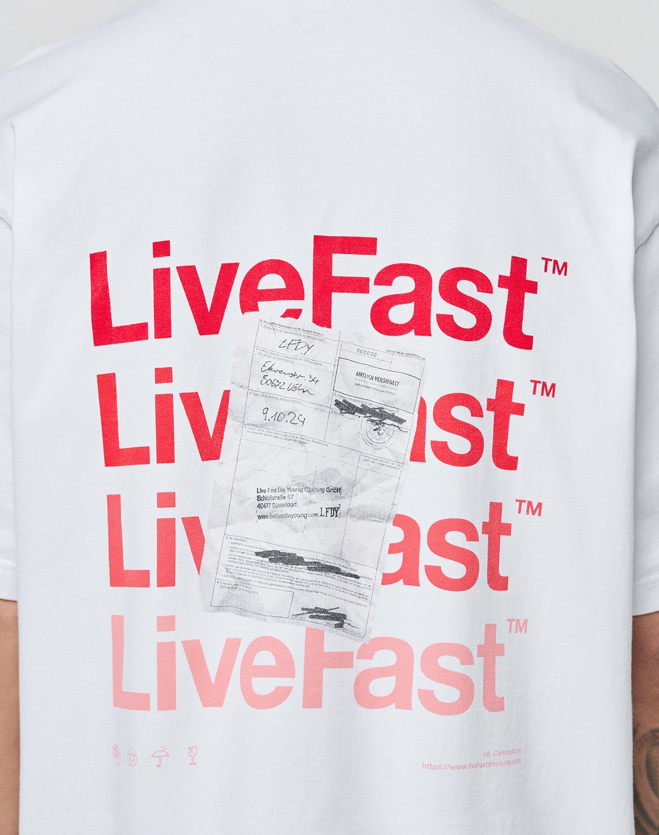 A person wears the LIVE FAST DIE YOUNG Basic Evidence Tee, a white 100% Baumwolle oversize T-shirt featuring bold Live Fast print on the back, faded receipt graphic, and striking front and back prints.