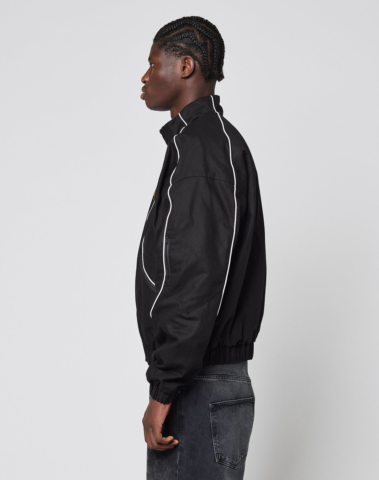 A person with braided hair stands in profile wearing the black LFDY Canvas Jacket from LIVE FAST DIE YOUNG, featuring white piping, multi-logo embroidery, and elastic cuffs, styled with dark jeans against a plain light gray background.