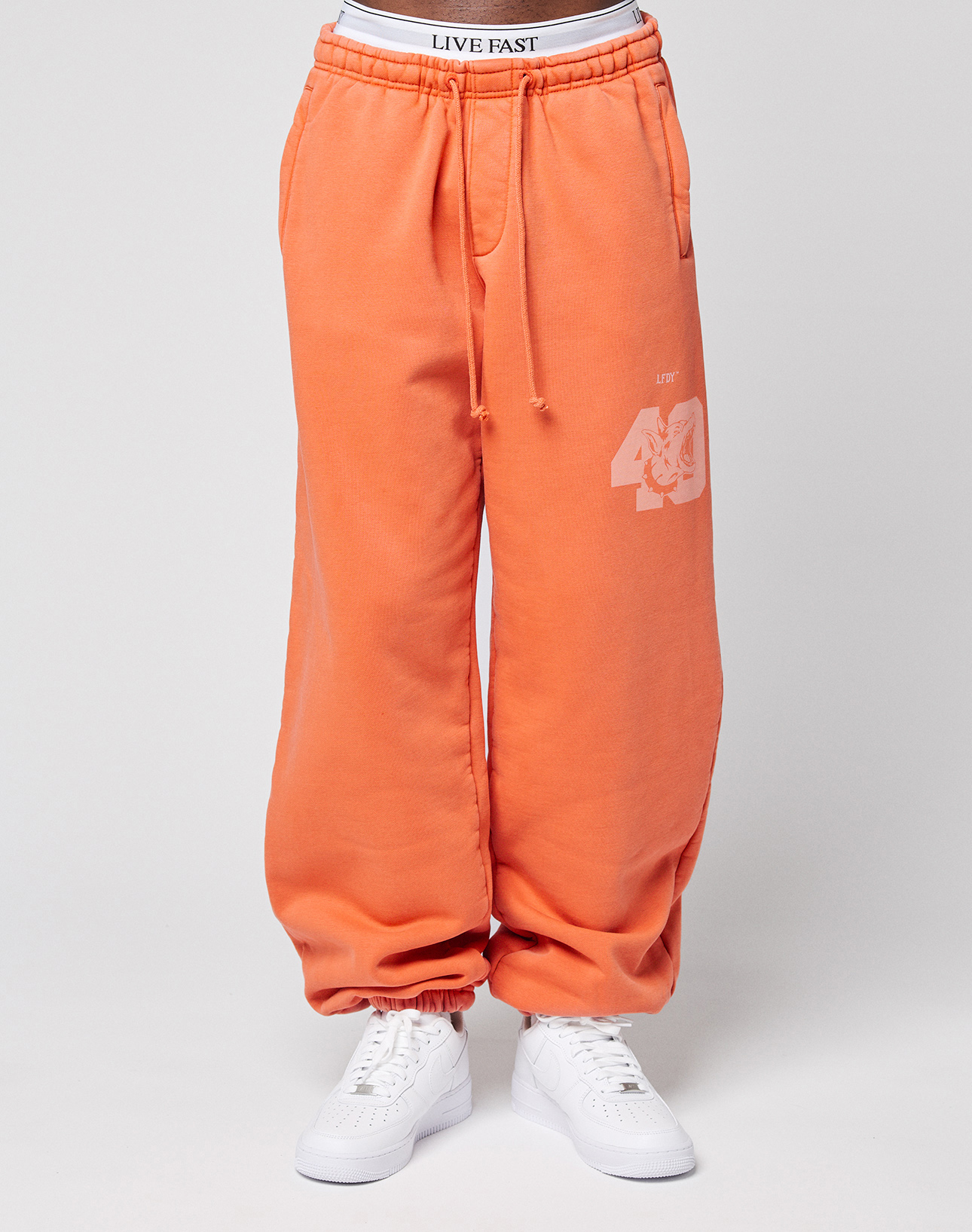 A person wears LFDY 40 Wide Cuff Jogger by LIVE FAST DIE YOUNG in orange with ‘40’ and print on the leg, white waistband with LIVE FAST text, side pockets, and white sneakers, standing against a light background.