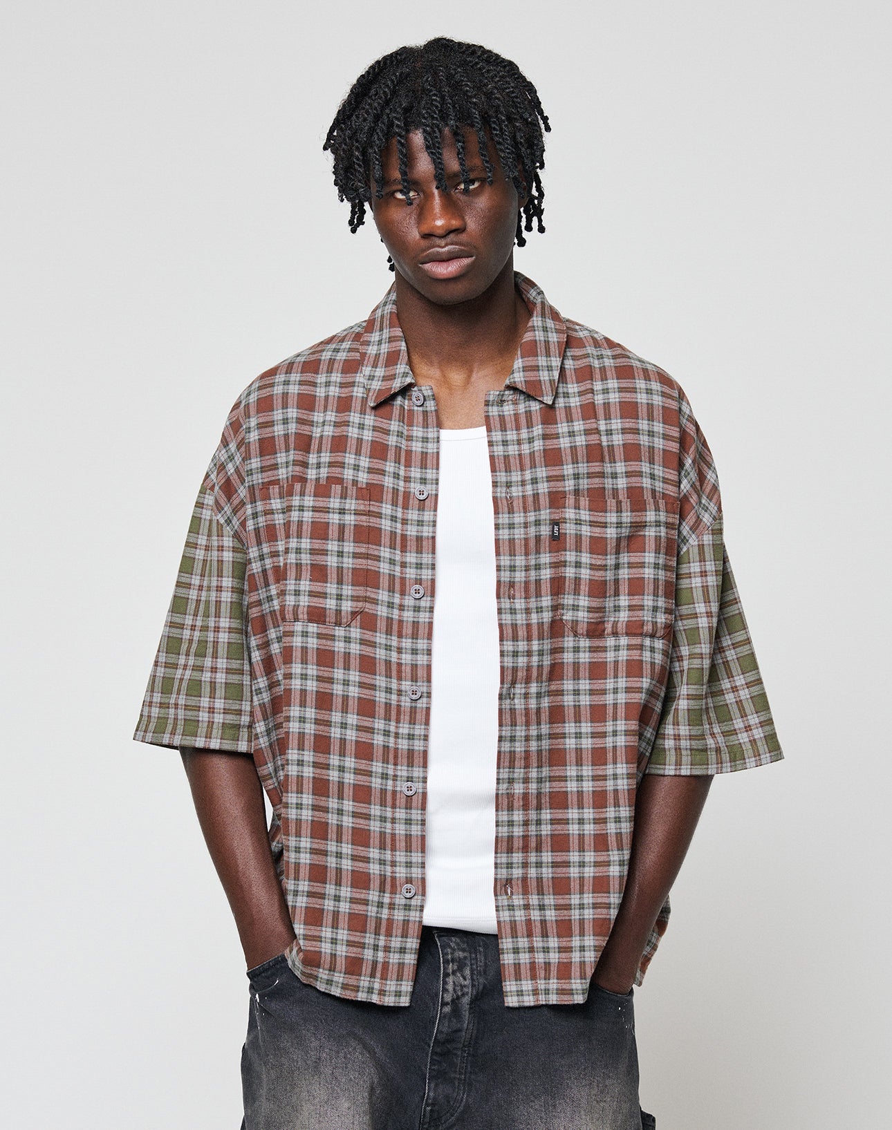 A young man with short dreadlocks wears the LIVE FAST DIE YOUNG LFDY Light Flannel—an oversized brown and green plaid shirt—over a white tank top and dark jeans, standing against a plain light-grey background.