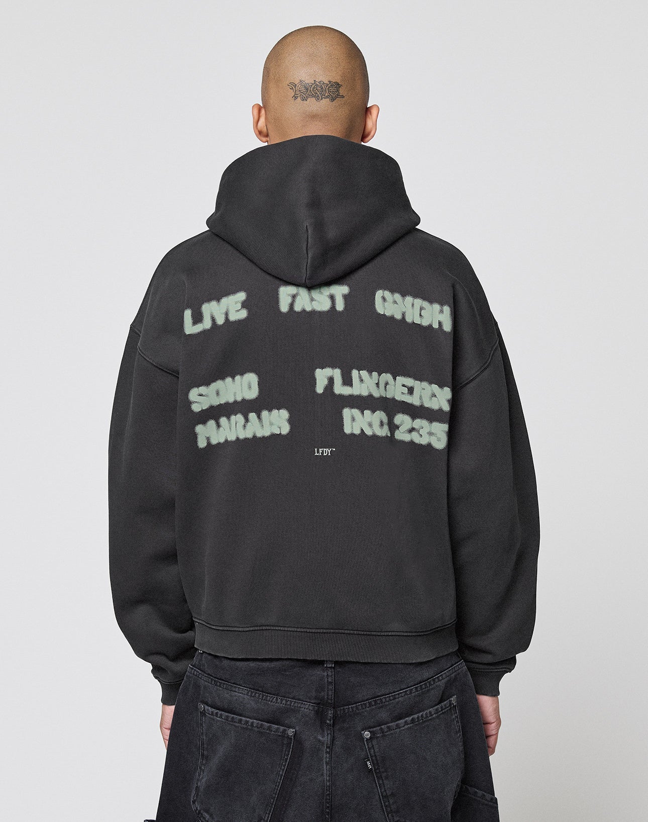 A person with a shaved head and tattoo wears the LIVE FAST DIE YOUNG Live Fast District Hooded Zip in black, featuring bold white spray-paint style text on the back for a signature streetwear look.
