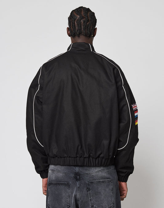 A person with braided hair, seen from behind, wears the black LFDY Canvas Jacket by LIVE FAST DIE YOUNG, featuring white piping and multi-logo patches including a UK flag and rainbow stripes on the sleeve. Faded black jeans complete the look. Plain white background.