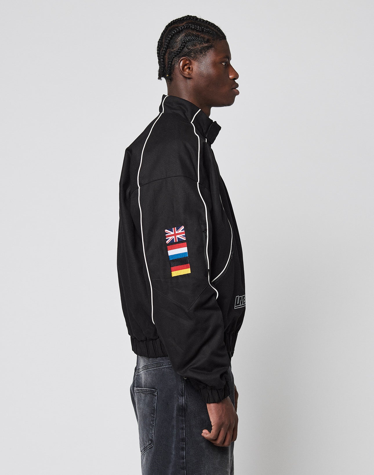A person in profile wears the LFDY Canvas Jacket by LIVE FAST DIE YOUNG—black with white piping, multi-logo patches and flag details, plus elastic cuffs. They have braided hair and pair it with faded black jeans.