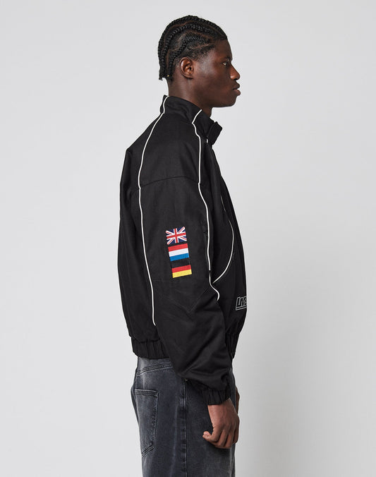 A person in profile wears the LFDY Canvas Jacket by LIVE FAST DIE YOUNG—black with white piping, multi-logo patches and flag details, plus elastic cuffs. They have braided hair and pair it with faded black jeans.