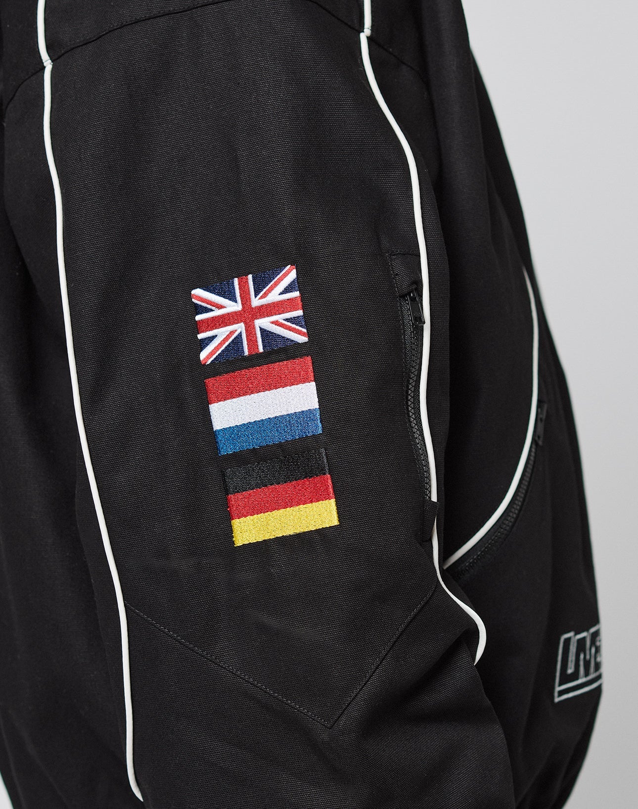 The LFDY Canvas Jacket by LIVE FAST DIE YOUNG is a black racing jacket with white piping, multicolor logo embroidery of the UK, France, Estonia, and Germany on the sleeve next to a zip pocket and sleek stand-up collar.