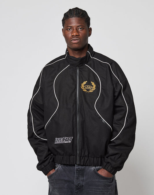 A man with braided hair wears the black LFDY Canvas Jacket by LIVE FAST DIE YOUNG, featuring white piping, multiple logo embroideries including a gold crest emblem, and LIVE FAST text near the hem. He stands with hands in his pockets against a plain background.