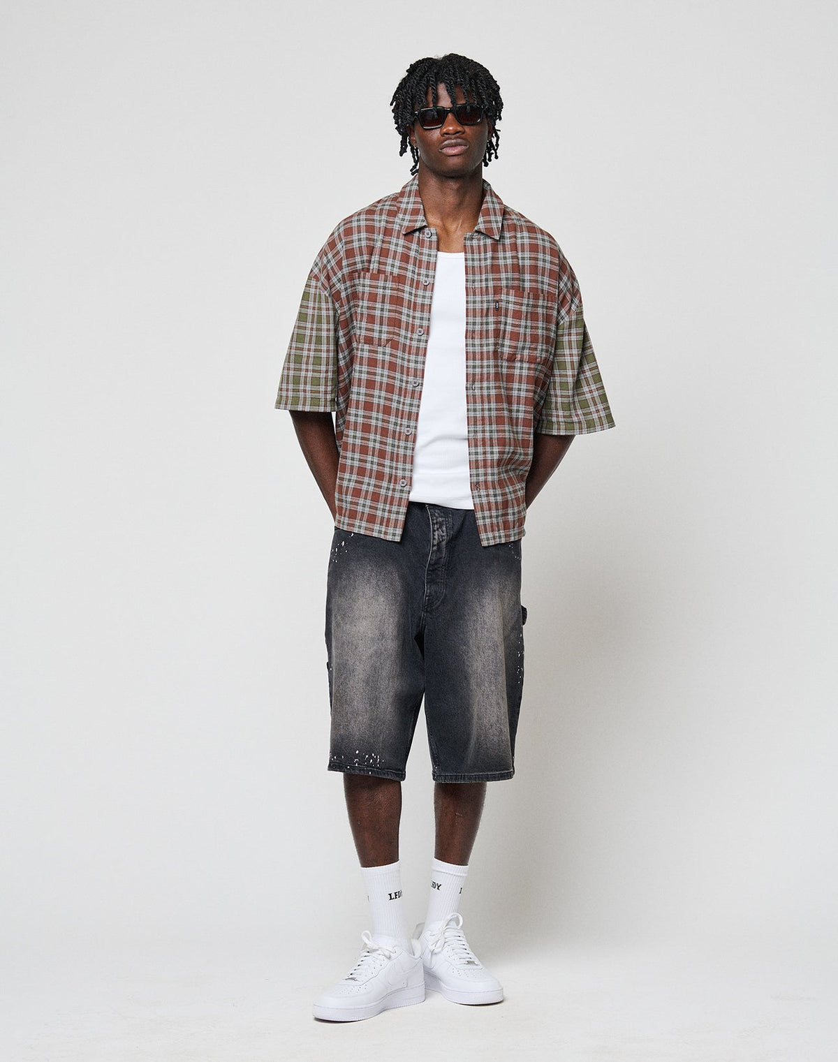 A man with short dreadlocks and sunglasses wears the LIVE FAST DIE YOUNG LFDY Light Flannel shirt over a white tee, paired with knee-length faded black denim shorts, white socks, and sneakers, standing against a plain light background.