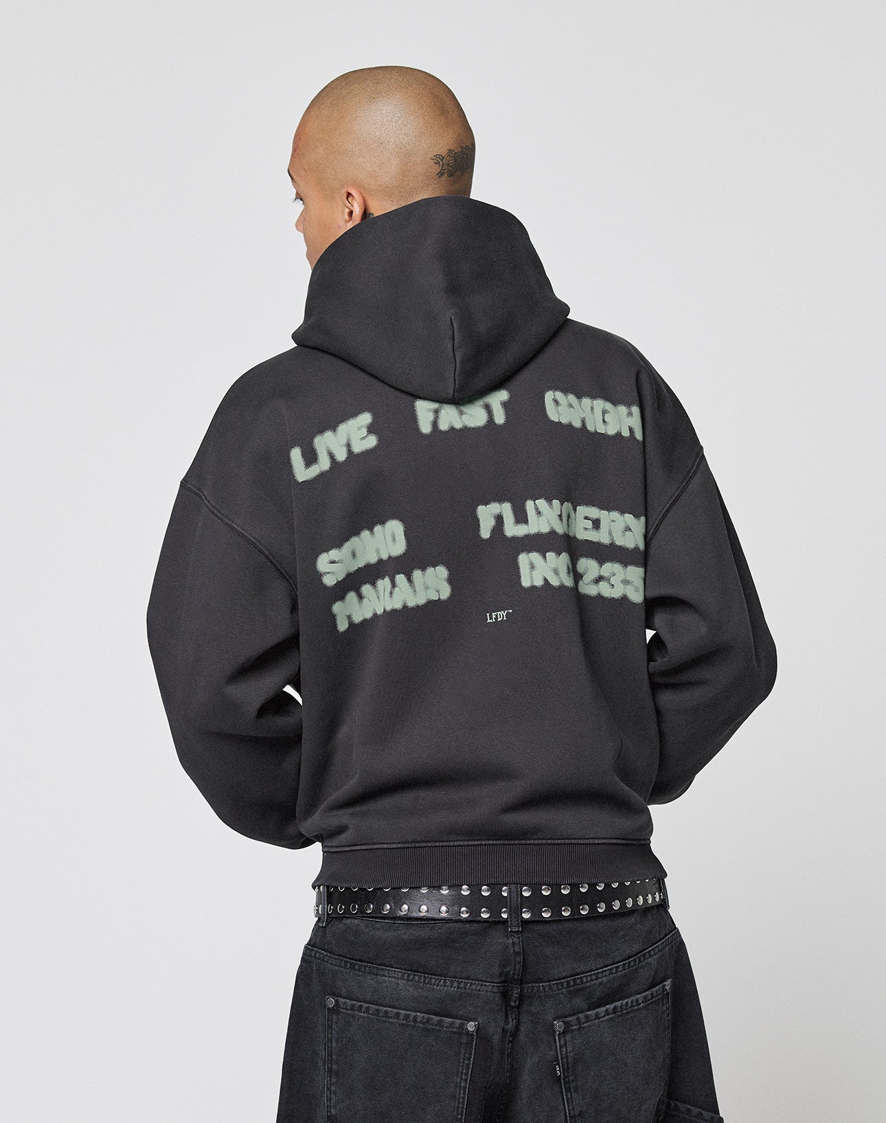 A person faces away wearing the LIVE FAST DIE YOUNG Live Fast District Hooded Zip with graffiti-style text, paired with black jeans and a studded belt for an effortlessly cool look.