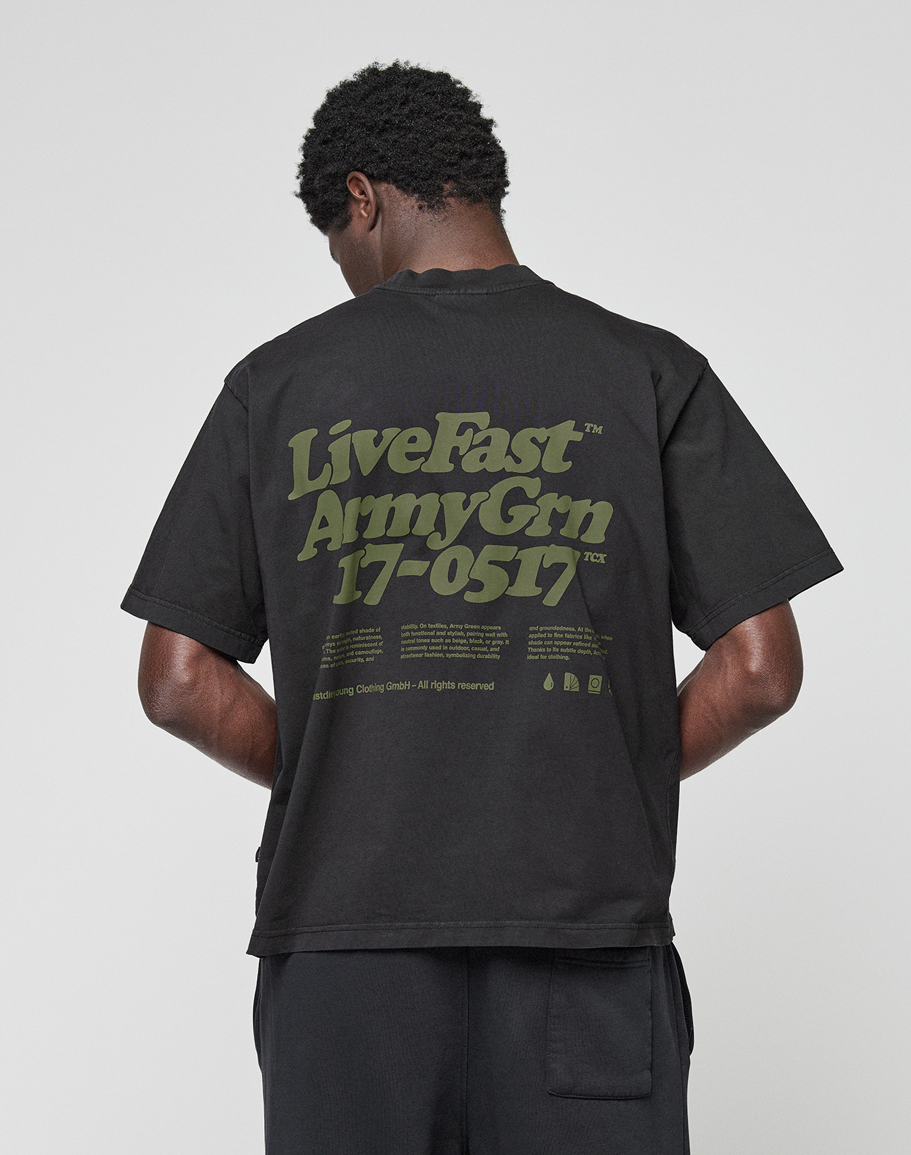 A person faces away from the camera wearing the LIVE FAST DIE YOUNG Basic 210 Tee, an oversized black 100% Baumwolle shirt featuring green “LiveFast ArmyGrn 17-0517” text on the back, against a plain light gray background.