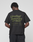 A person faces away from the camera wearing the LIVE FAST DIE YOUNG Basic 210 Tee, an oversized black 100% Baumwolle shirt featuring green “LiveFast ArmyGrn 17-0517” text on the back, against a plain light gray background.