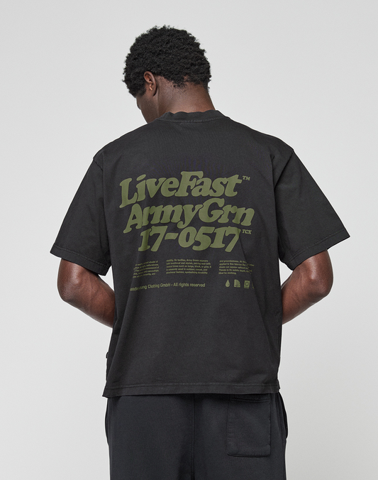 A person faces away from the camera wearing the LIVE FAST DIE YOUNG Basic 210 Tee, an oversized black 100% Baumwolle shirt featuring green “LiveFast ArmyGrn 17-0517” text on the back, against a plain light gray background.