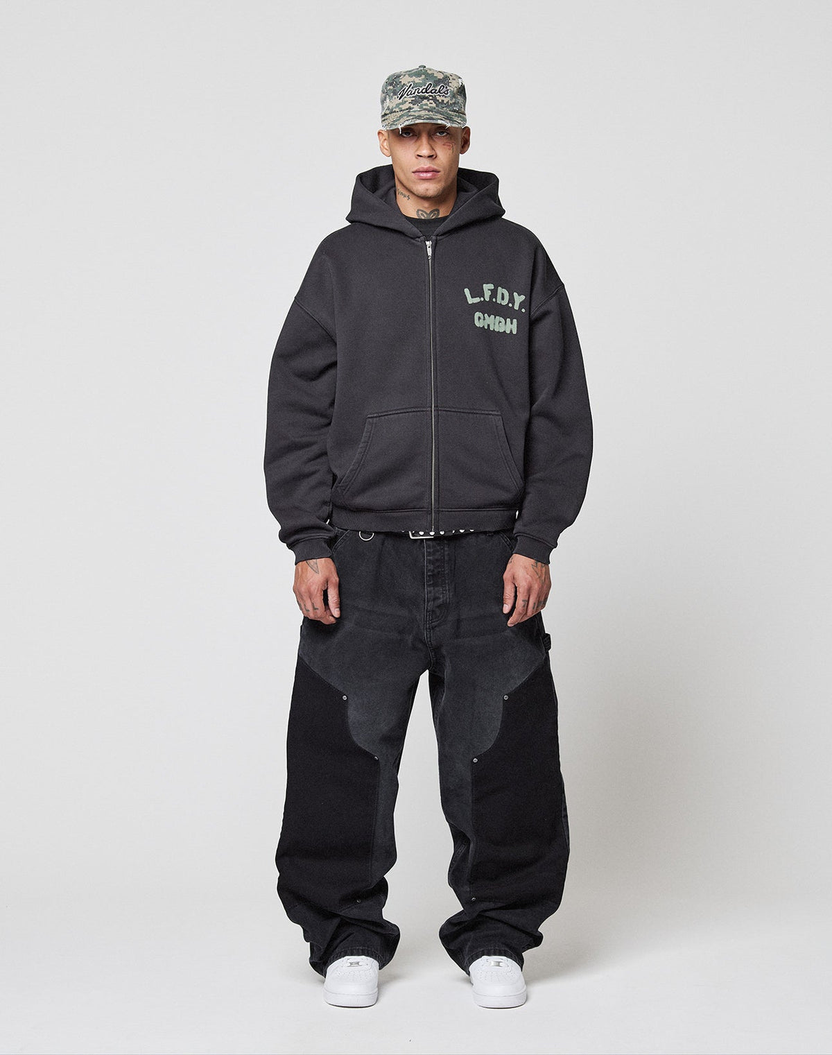 A man in a gray camo cap, the Live Fast District Hooded Zip by LIVE FAST DIE YOUNG (black, branded chest), loose black jeans with Wasch-Effekt panels, a black belt, and white sneakers stands against a plain white background.