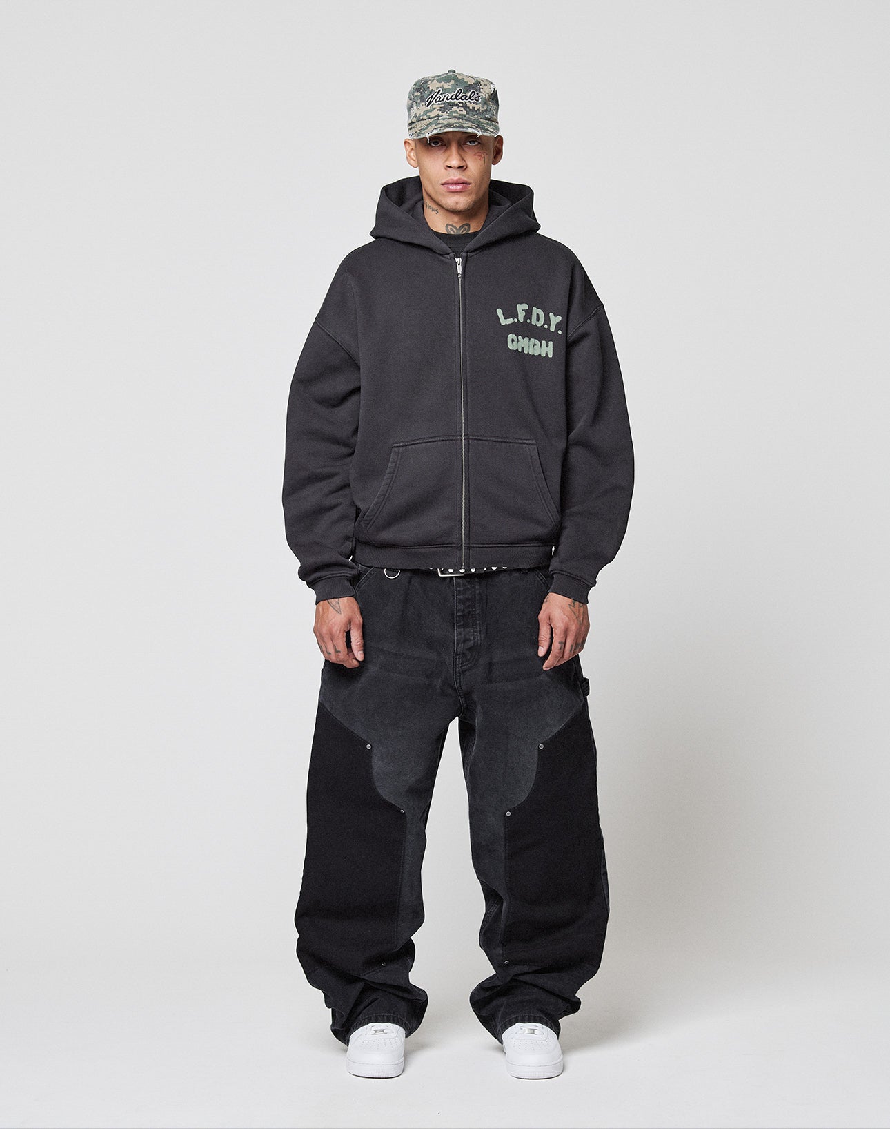 A man in a gray camo cap, the Live Fast District Hooded Zip by LIVE FAST DIE YOUNG (black, branded chest), loose black jeans with Wasch-Effekt panels, a black belt, and white sneakers stands against a plain white background.