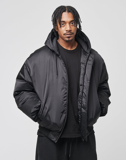 A man with braided hair wears a black LIVE FAST DIE YOUNG LFDY Winter Bomber Jacket with Kapuze and black pants, standing with hands in his pockets and looking at the camera against a plain light background.