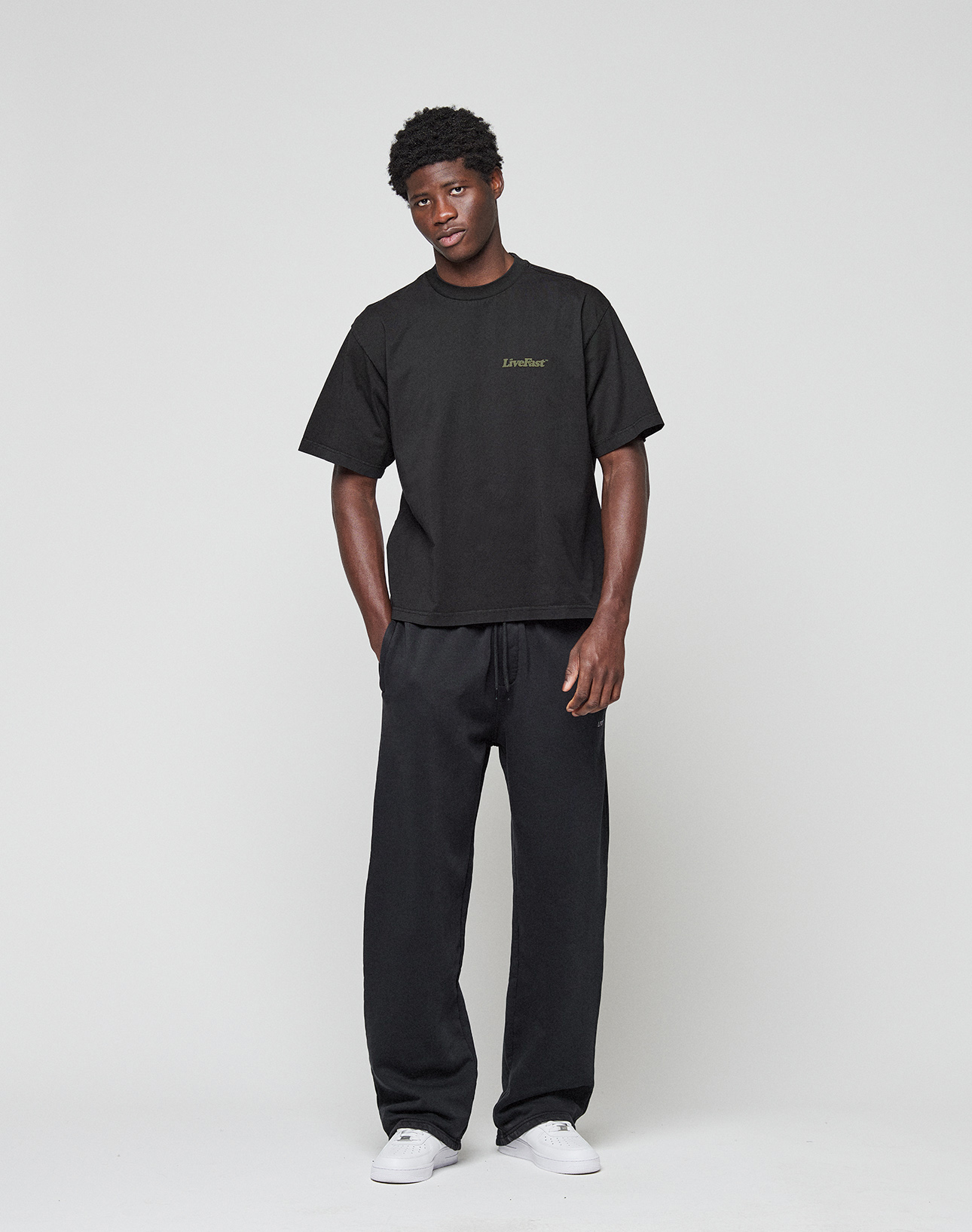 A man with short curly hair and a neutral expression stands against a white background wearing the LIVE FAST DIE YOUNG Basic 210 Tee, an oversize fit black t-shirt made from 100% Baumwolle, black drawstring pants, and white sneakers.