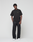 A man with short curly hair and a neutral expression stands against a white background wearing the LIVE FAST DIE YOUNG Basic 210 Tee, an oversize fit black t-shirt made from 100% Baumwolle, black drawstring pants, and white sneakers.