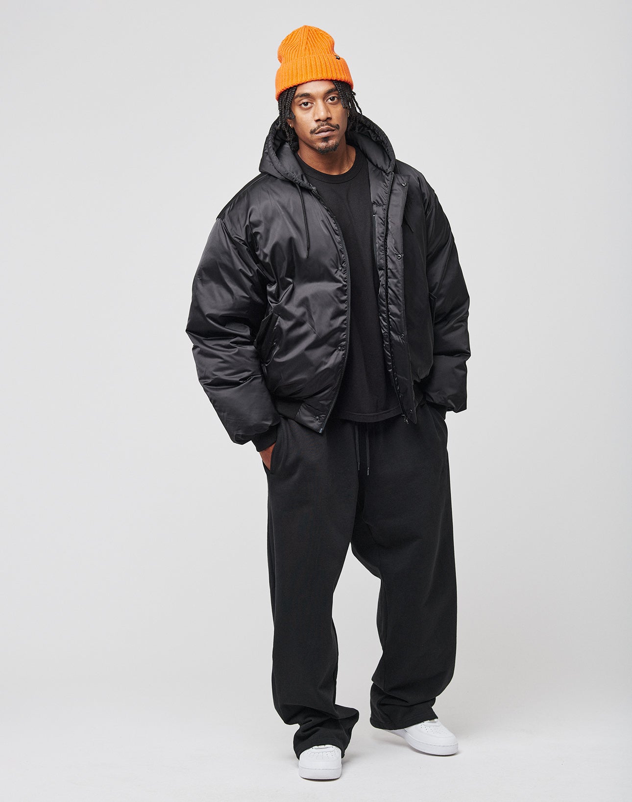 A man stands against a plain background wearing the LIVE FAST DIE YOUNG LFDY Winter Bomber Jacket, loose black pants, a black t-shirt, white sneakers, and an orange knit beanie, looking directly at the camera with a neutral expression.