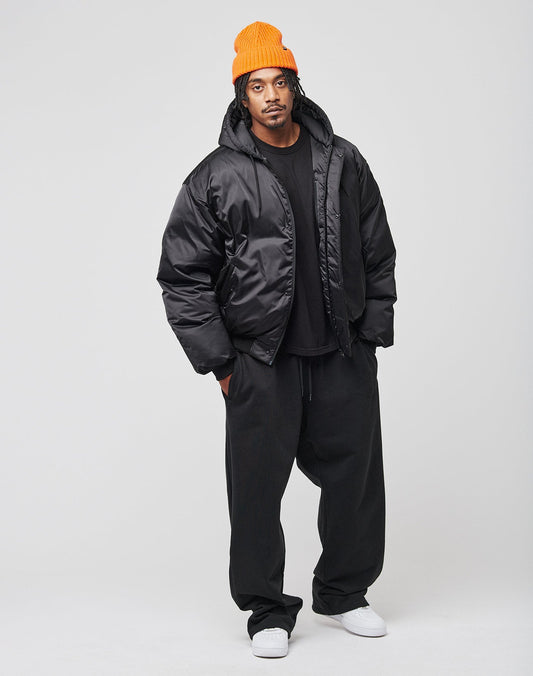 A man stands against a plain background wearing the LIVE FAST DIE YOUNG LFDY Winter Bomber Jacket, loose black pants, a black t-shirt, white sneakers, and an orange knit beanie, looking directly at the camera with a neutral expression.