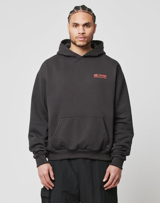 A man with braided hair wears the LIVE FAST DIE YOUNG Hit and Run Hooded, an oversized black hoodie with a red and white discharge logo on the chest, standing against a plain white background.