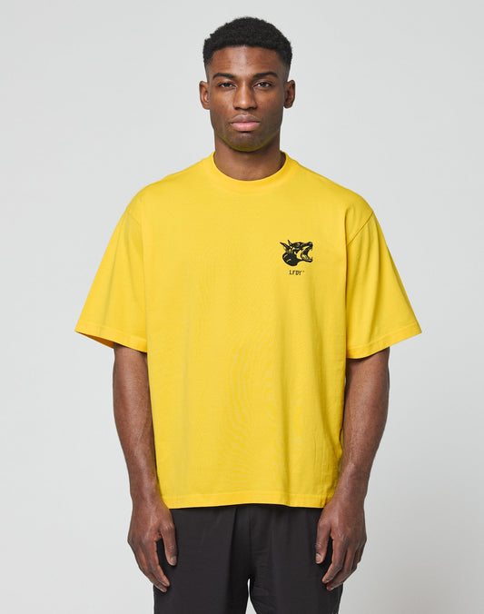 A man stands facing forward wearing the LFDY Circle Dog Tee by LIVE FAST DIE YOUNG, a loose-fitting yellow oversize t-shirt with a small black LFYT graphic on the upper left chest, made from 100% cotton, paired with black pants against a light gray background.