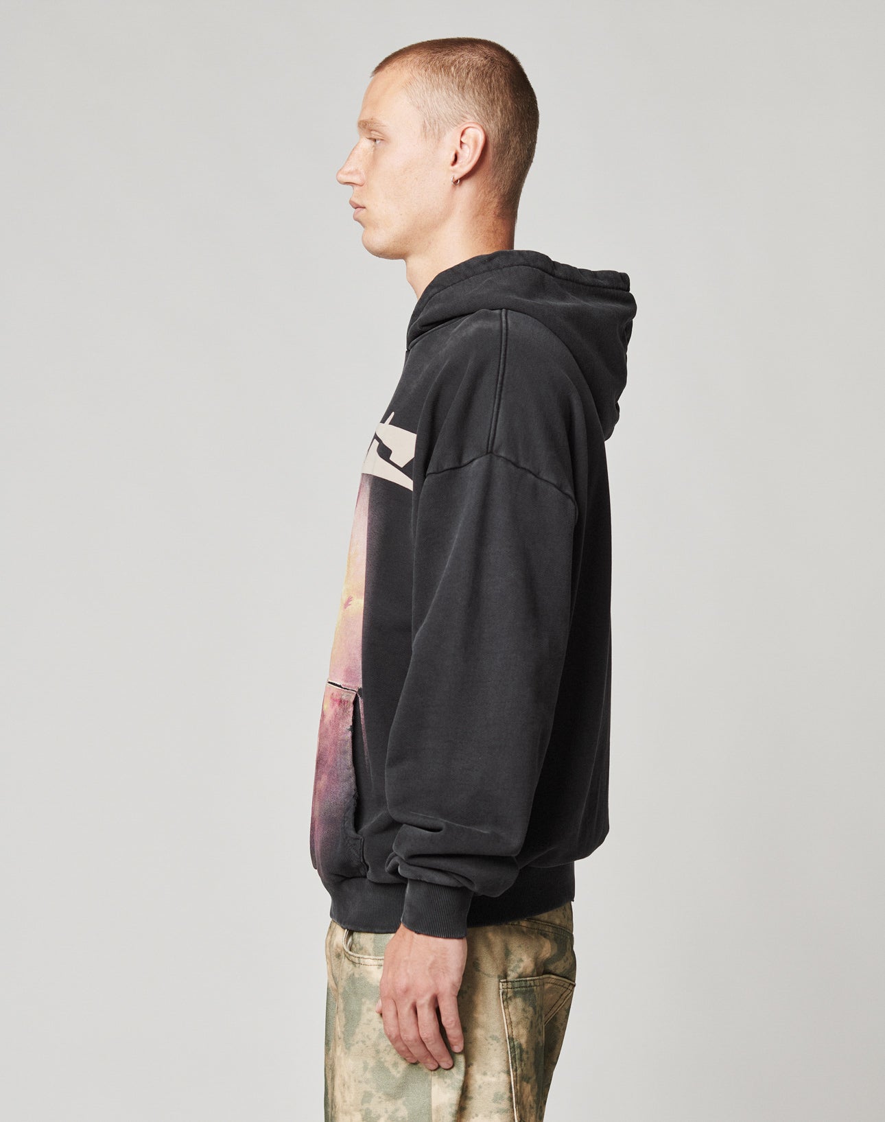 A person with short hair stands in profile, wearing the LIVE FAST DIE YOUNG LFDY Angel Hooded black sweatshirt with a graphic design and light camouflage pants against a plain gray background.