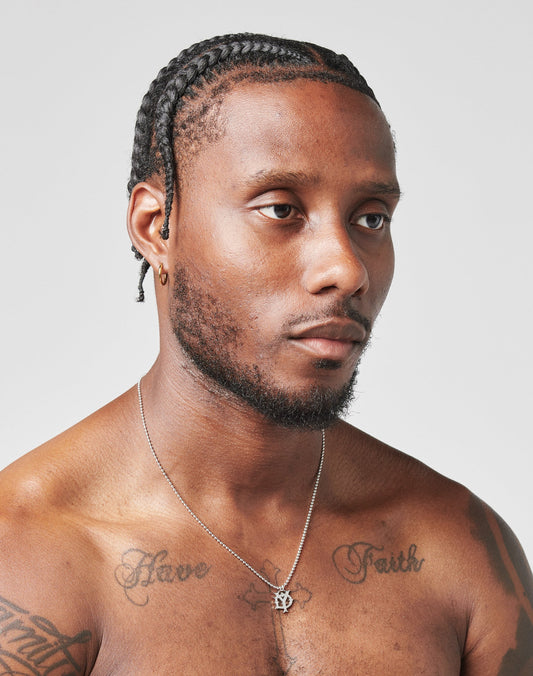 A man with braided hair and a close-trimmed beard glances sideways. He wears the LIVE FAST DIE YOUNG DY Chain necklace with adjustable length and zirconia stones. His chest tattoos read Have and Faith. The background is plain and light-colored.