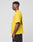 A person in profile wears the LIVE FAST DIE YOUNG LFDY Circle Dog Tee, a bright yellow 100% Baumwolle t-shirt with a back graphic, and black shorts, standing against a plain light gray background.